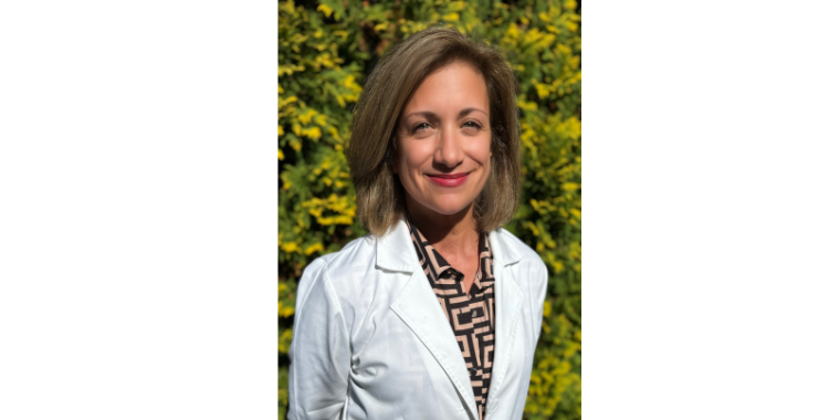 Dr. Narr stands in front of green plants and flowers in her white coat. 